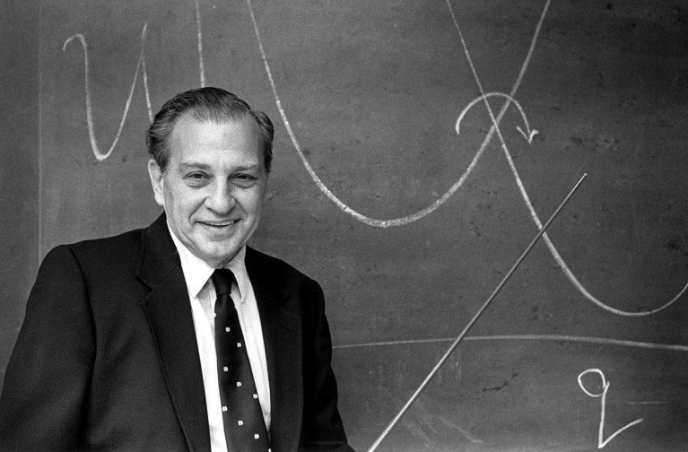 Portrait of Rudolph Marcus smiling in front of a classroom chalkboard displaying handwritten notes.