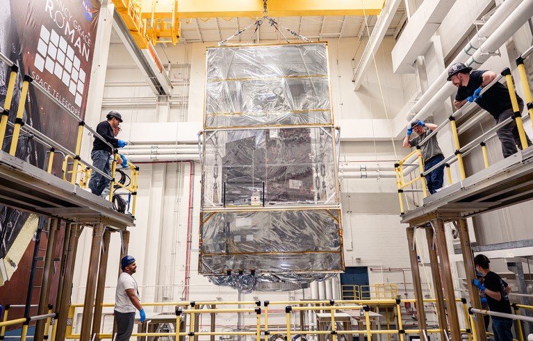 A large, rectangular satellite component being carefully lifted by a yellow overhead crane inside a spacious industrial facility. Four technicians wearing helmets observe and guide the process from elevated platforms, with a banner labeled "Roman Space Telescope" visible on the left side.