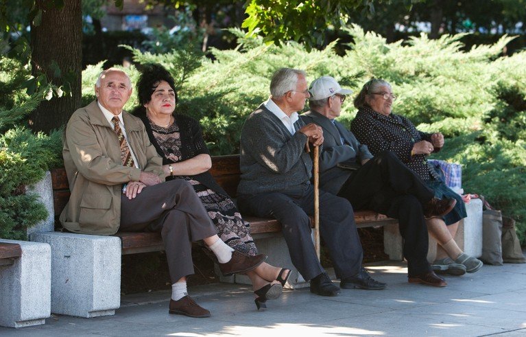 Five elderly people sitting on a bench in Yuzhen Park, Sofia, Bulgaria, surrounded by greenery and shaded by trees. The group includes three men and two women dressed in formal and casual attire, with one man holding a walking cane.