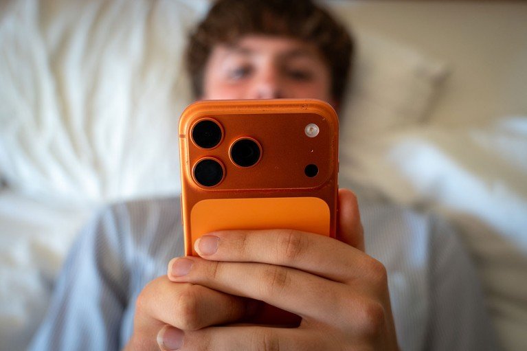 An anonymous teenage boy looks at a smartphone while lying in bed.