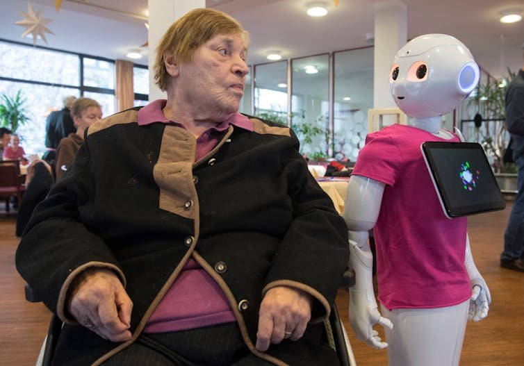 a seated elderly woman looks at a humanoid robot that has turned its face toward her