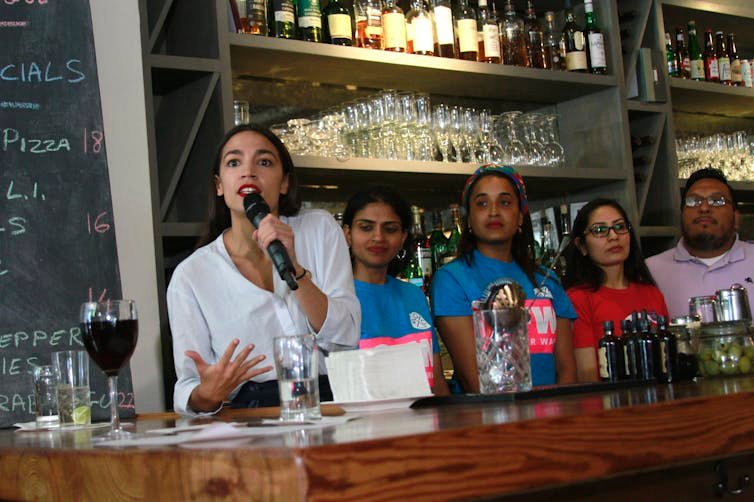 U.S. Rep. Alexandria Ocasio-Cortez speaks into a microphone behind a bar alongside four other people.
