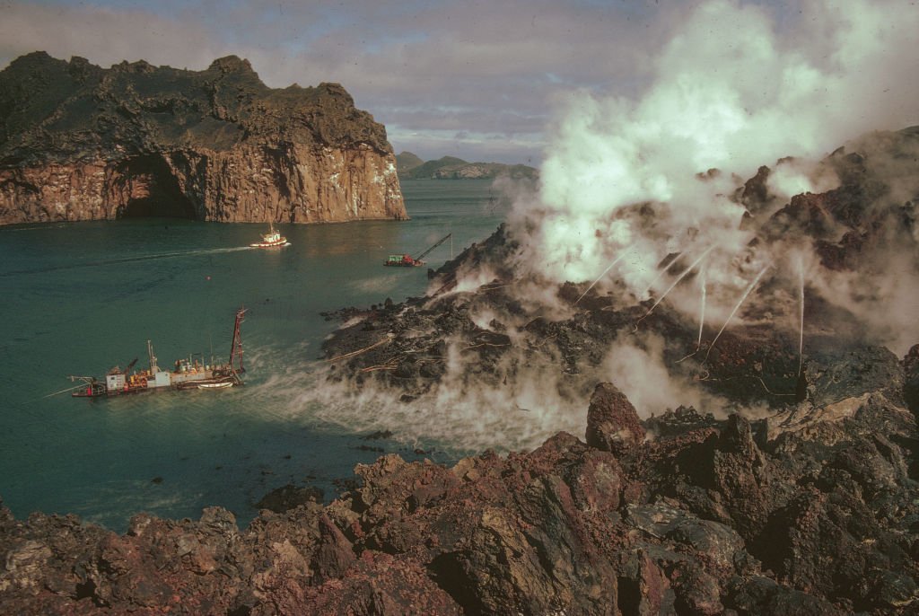 Large landscape image of rocky island with water hoses blasting water onto steaming black lava flows. Several ships can be seen in the water as well as other rocky islands and outcroppings.