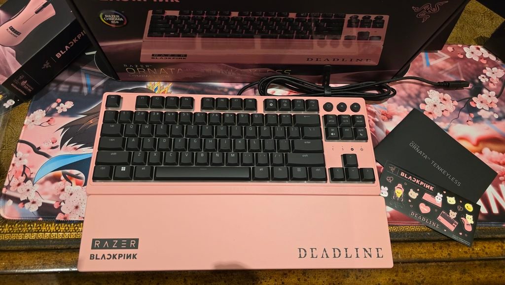 Top down image of the Razer BLACKPINK Ornata keyboard with a sticker sheet from Razer BlackPINK. There are several stickers on there with "Deadline" and Razer pink peripherials.