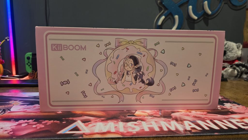 Outer "KiiBOOM" box showing a kawaii cartoon girl inside a donut that has a bow on top of the donut. It's a pastel pink colour palette with candy around the edges in pastel. She is wearing glasses and has her hair in pigtails with one side pink and one side brown like neopolitan icecream.
