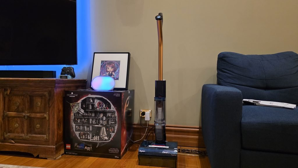 The Dyson unit sitting between a couch and a large LEGO box in the lounge room. It's plugged in, turned on, and sitting in its caddy charging.