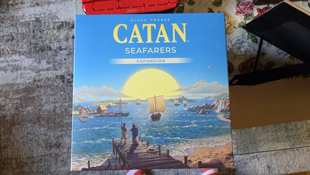 Settlers of Catan box showing the "expansion" and a picture of boats the water as two people stand on the pier and watch.