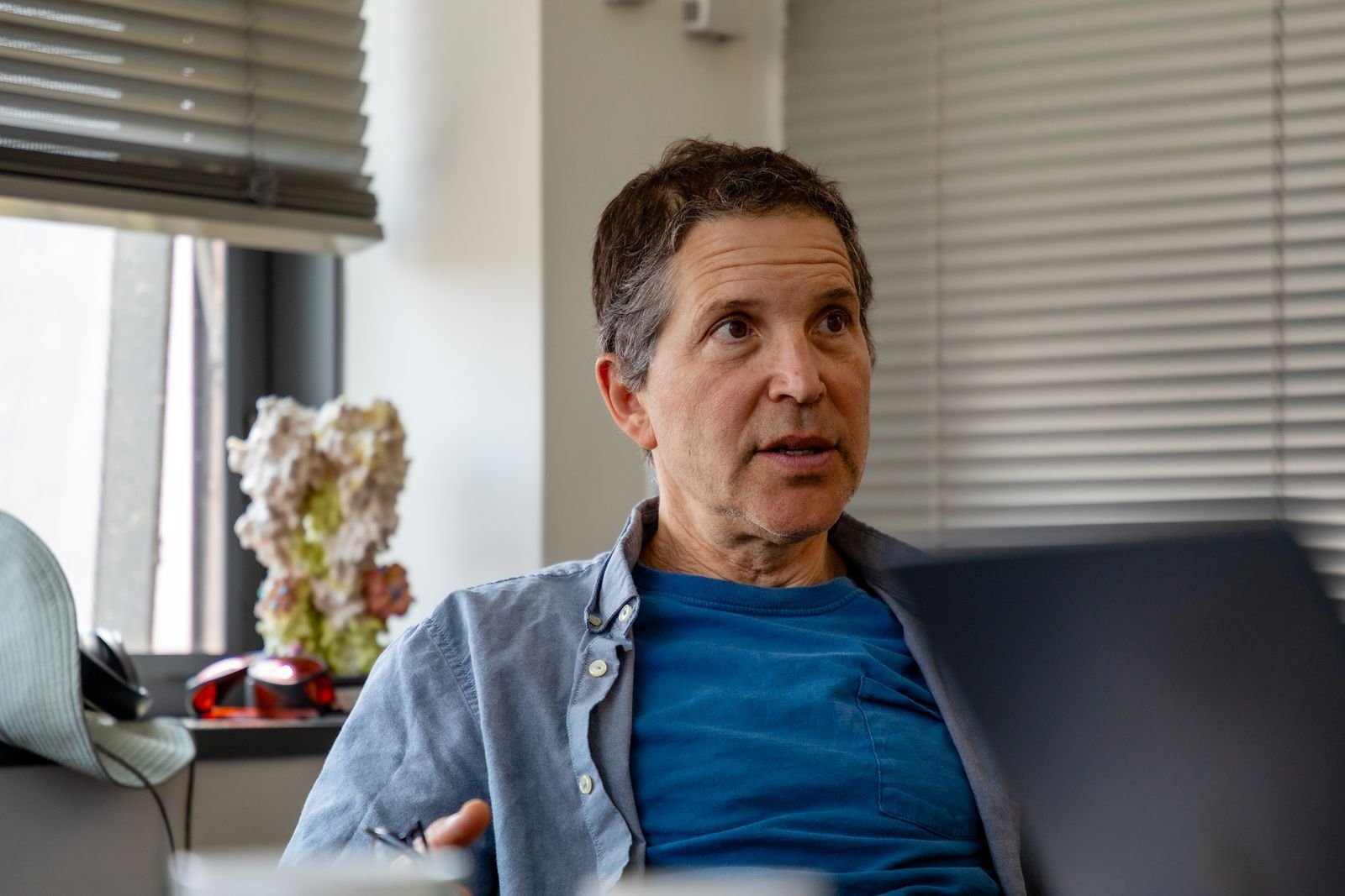 A white man with short dark hair and a blue shirt sits in the corner of an office and looks off camera. He looks to be...