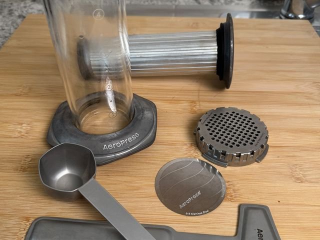 AeroPress Premium Coffee Press on a wooden cutting board beside scattered accessories and parts.