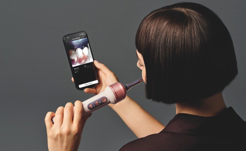 A person using the pink Dyson CameraJet toothbrush while holding a phone showing a live view of their teeth.