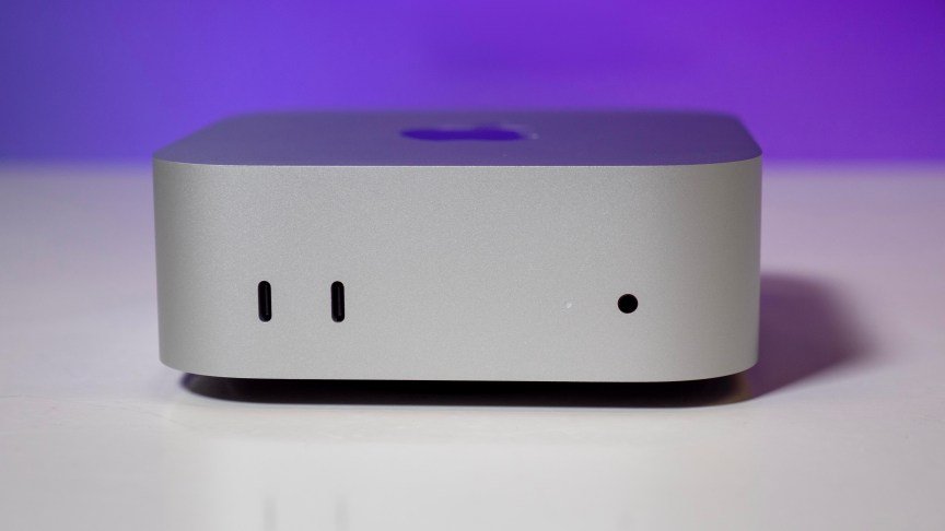 the front view of an apple mac mini sitting on a glossy white table in front of a purple background. 