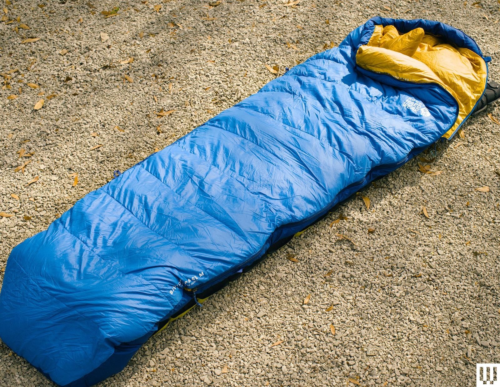 Long blue sleeping bag with yellow interior laid out on top of tan gravel surface