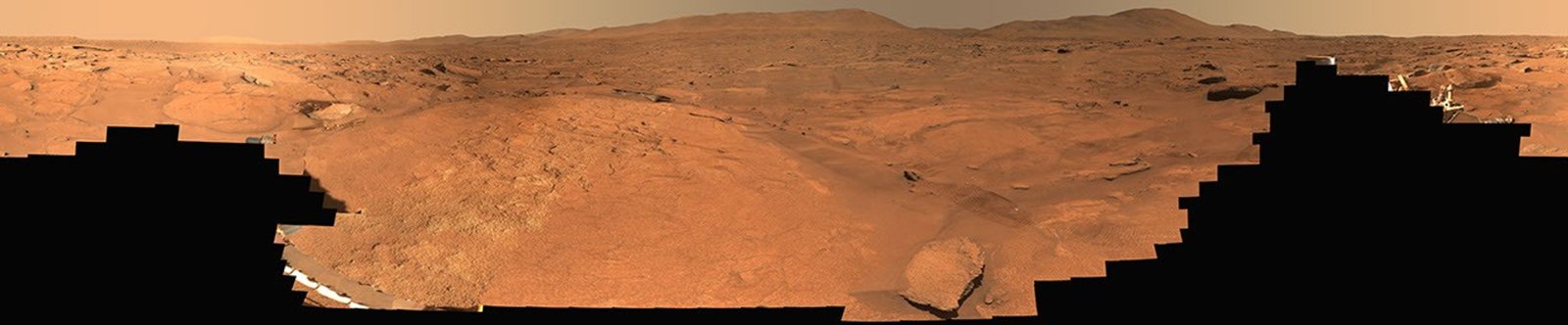 A panoramic view of a barren, reddish-orange Martian landscape stretching toward distant hills, partially obscured by the jagged black silhouettes of a rover in the foreground.