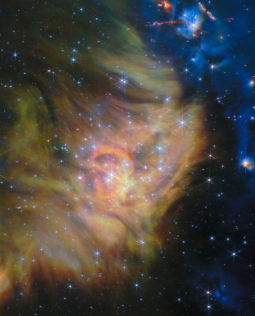 A star-forming region. The left two thirds of the image is blanketed in thick clouds of green and yellow gas and dust that form swirling patterns. Some of the gas is spread into thin, thready wisps. The right third of the image is the black background speckled with stars and multi-colored dots that represent distant galaxies. Bright stars are scattered throughout the clouds, with the densest cluster situated in the center of the scene. Each is crowned with six long spikes and two shorter spikes created by the optics of the telescope’s mirrors. In the upper right, a couple of hidden stars blast out long, glowing jets of material that glow in blue.