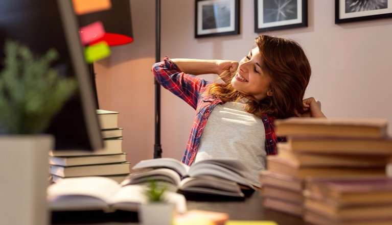 A woman sitting at her home desk smiles and stretches her arms above her head. She is surrounded by stacks of books, a few are open in front of her and her computer.