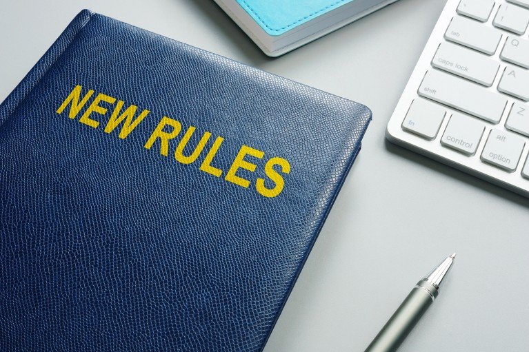 Close up of a hardback book with the title "NEW RULES" resting upon an office desk beside a pen and computer keyboard.