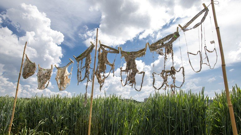 Various pieces of handmade netting or woven fabric hanging on a wooden frame outdoors against a cloudy sky and tall green grass in the background.