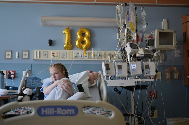 A young woman sits somberly in a hospital bed surrounded by medical equipment. Above her on the wall hands a bright gold birthday balloon in the shape of the number 18.