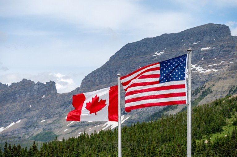 The Canadian and American flags wave side by side in Glacier National Park, with mountain terrain in the background.