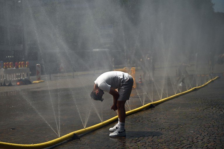 A man bends over to cool his head at a row of water sprinklers in a yellow hose spraying mist across a city street