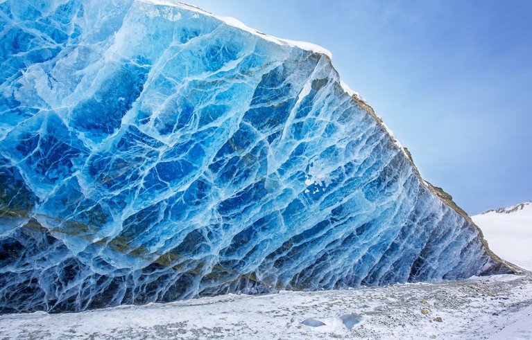 A glacier ice wall, showcasing vivid blue ice crossed by intricate white veins. Snow-covered foreground and pale blue sky highlight glacier scale.