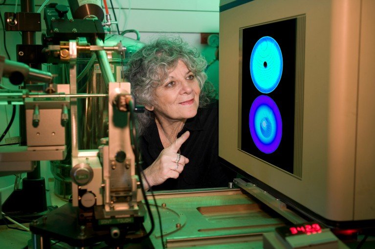 A portrait of Professor Ada Yonath in a laboratory pointing at a monitor displaying two blue circular imaging patterns, surrounded by x-ray diffraction equipment