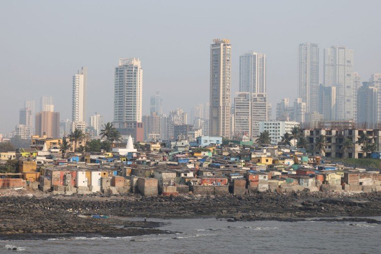 Low-rise slum housing is seen along a rocky waterfront, with tall modern skyscrapers towering behind in the background on a hazy day