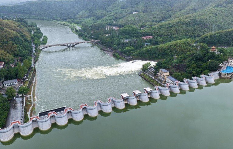 A large dam spanning a river surrounded by green hills and forested areas, with water flowing through spillways. Key features include a curved concrete structure with multiple gates, a downstream bridge, and buildings near the dam.