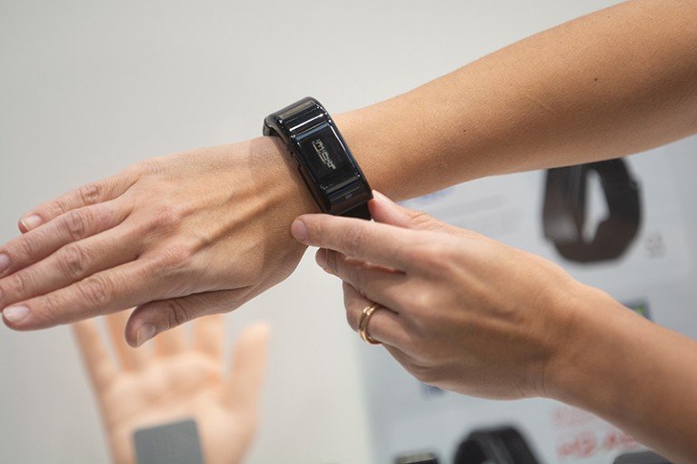 Close-up of forearm and hand with a black blood pressure monitor positioned on the wrist. A second hand is touching the monitor.