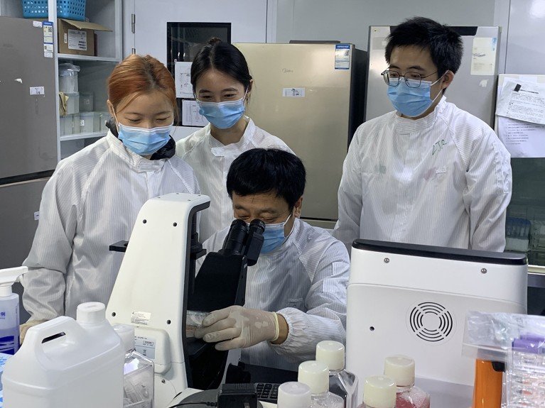 Hongkui Deng operates a microscope in a laboratory, while three researchers wearing face masks and cleanroom lab coats gather around him.