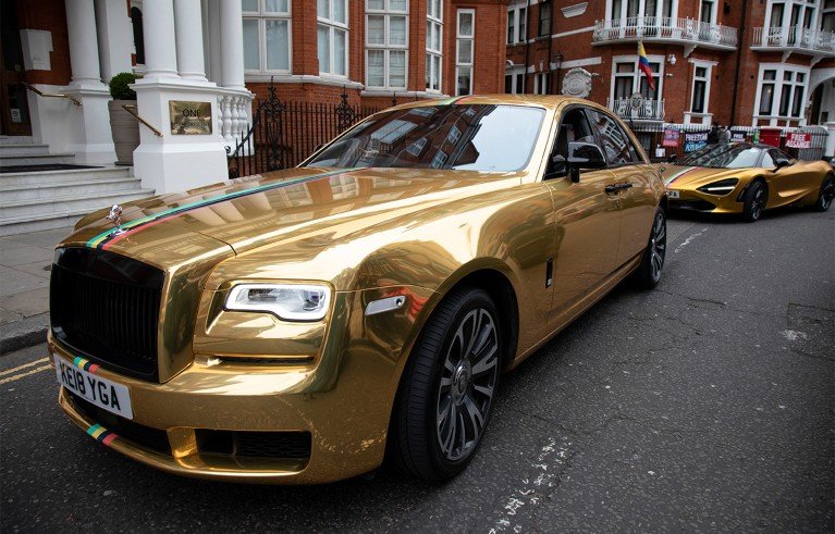 A gold Rolls-Royce parked on a street outside ornate red-brick townhouses.