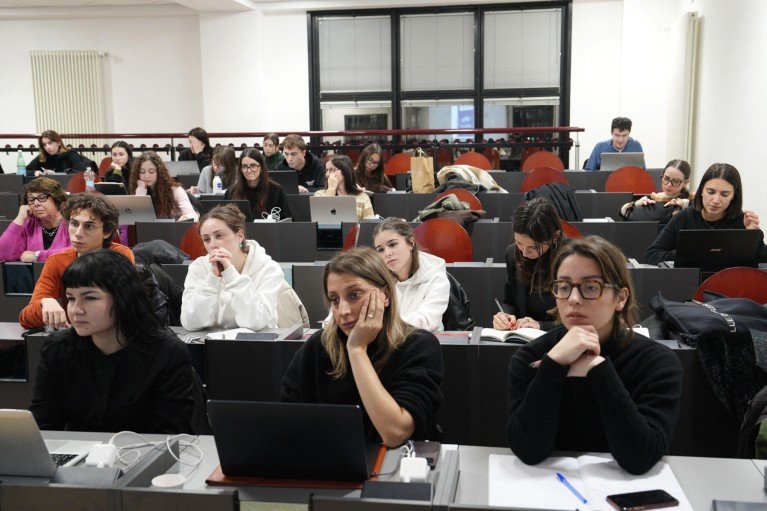 Students seated in a tiered lecture hall work on laptops and take notes.