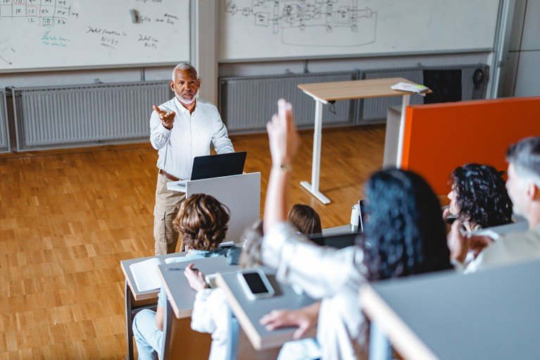 Professor at a lectern gestures towards attentive students in a tiered lecture hall; one student raises a hand. Whiteboards line the walls.