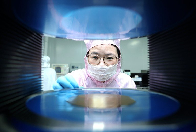 A female worker wearing PPE is seen through gap in a machine containing a semiconductor.