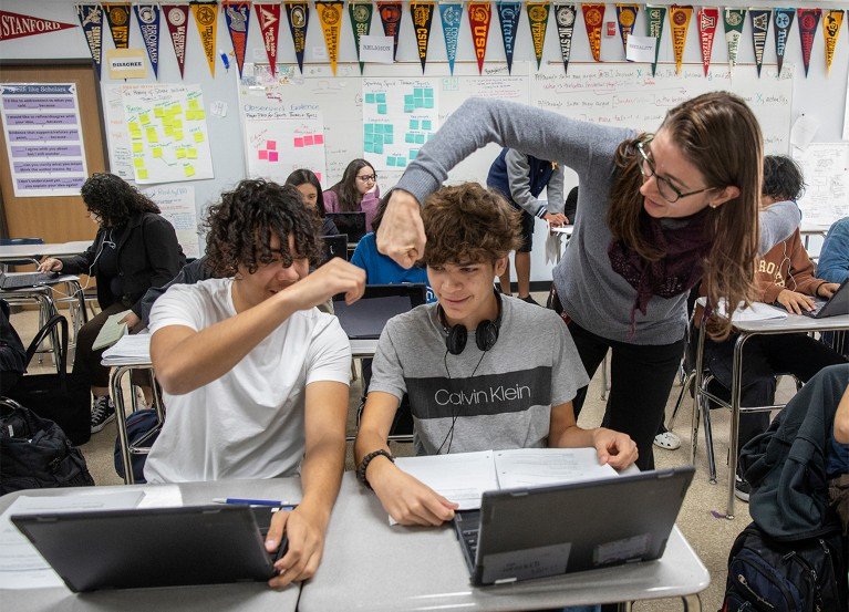 Seatec high-school student with a laptop receives congratulatory fist bump from teacher. Classroom walls display university pennants, posters and notes.
