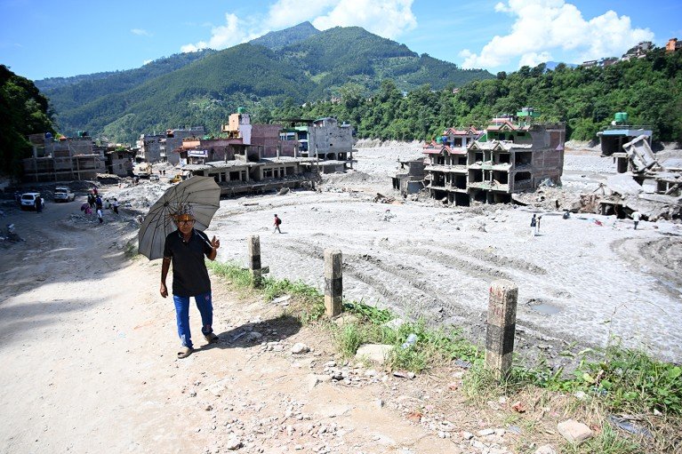 A man carrying a sun umbrella walks past muddy houses damaged by flash floods in Nepal, with green mountains rising in the background.