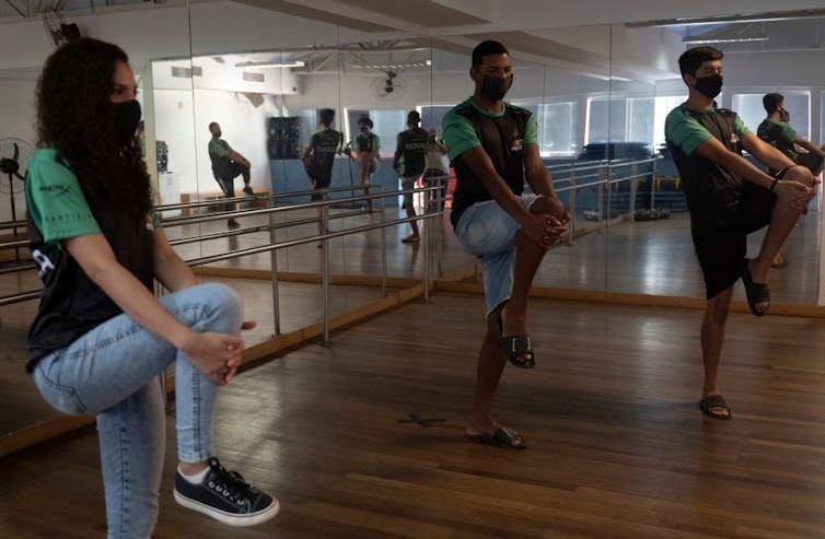 Two young men and one young woman balance on one leg while clasping the raised knee of their other leg with both hands.