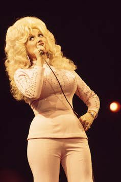 Young woman with billowing, blonde hair rolls her eyes while singing into a microphone, with one hand on her hip.