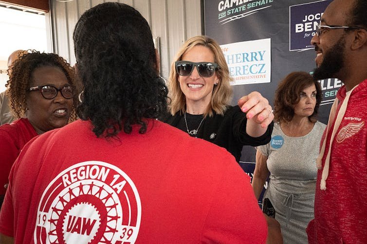 A woman wearing sunglasses smiles at a woman wearing a red t-shirt.