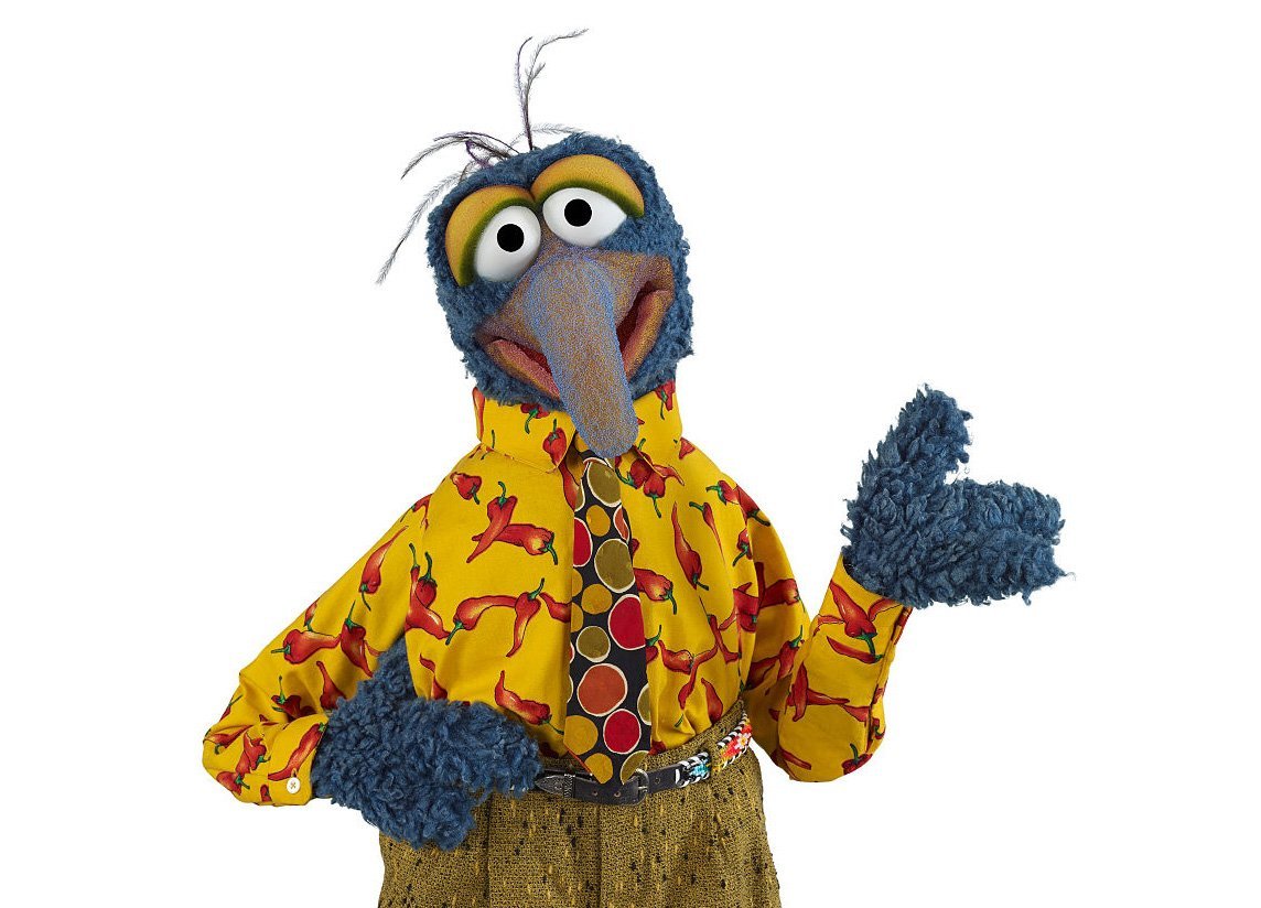 A puppet with big eyes, a long droopy nose looks at camera in a yellow button down with red peppers, a tie, and pants.