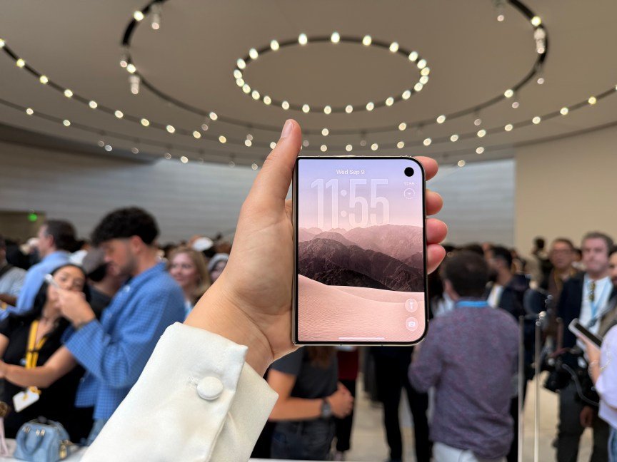 hand holding the iPhone Duo in a busy room, with the front screen showing mountains and the time 11:55