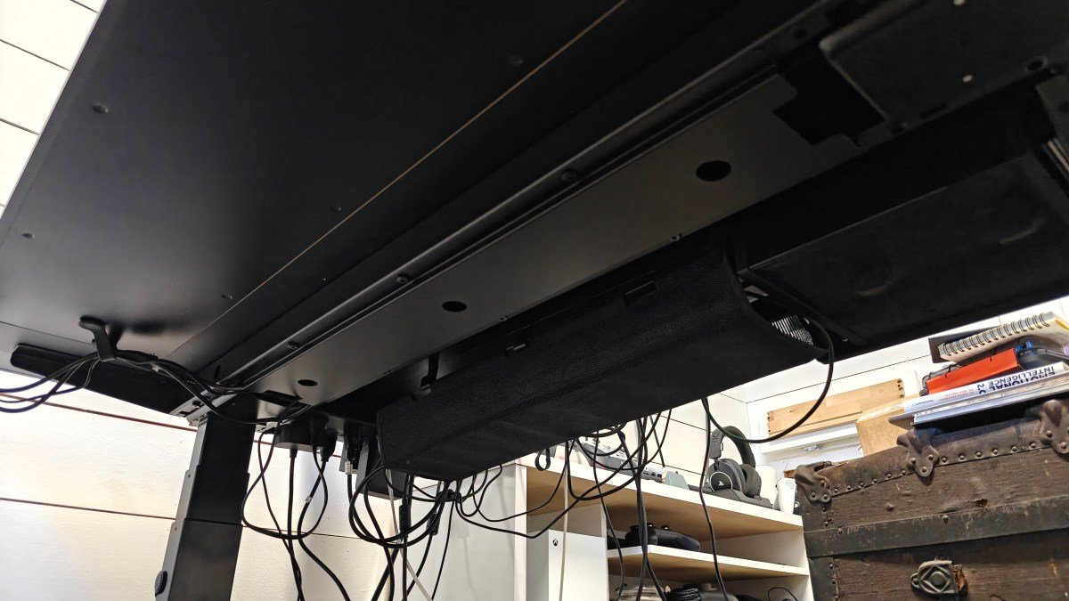 View underneath the MagVida standing desk with cables and a white shelf and brown chest in the background.
