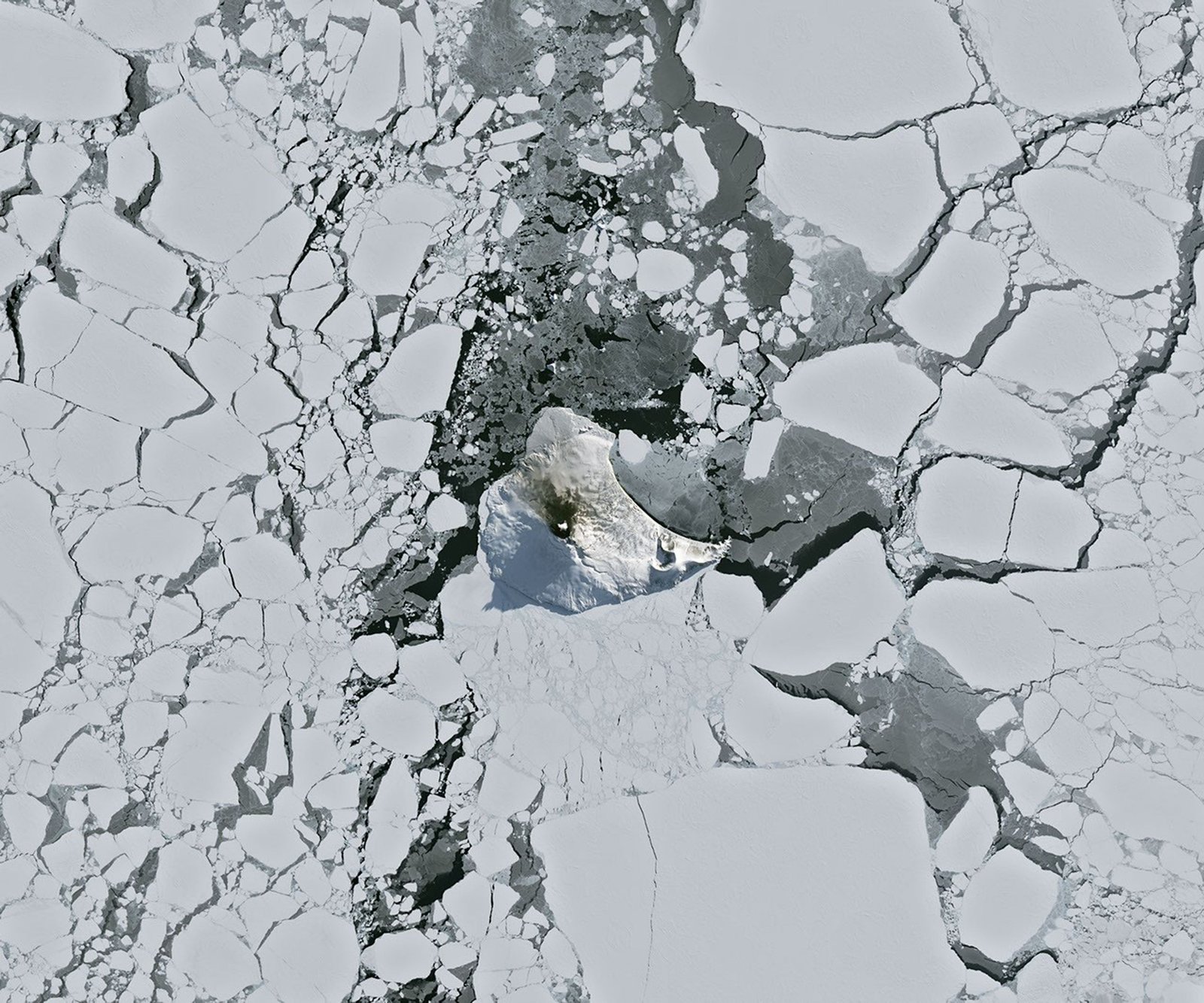 A small ice- and snow-covered island with an active volcanic crater at its center is surrounded by drifting pieces of sea ice. A thermal signal and small plume appear in the crater, and ash darkens the snow on the volcano’s northern slopes.
