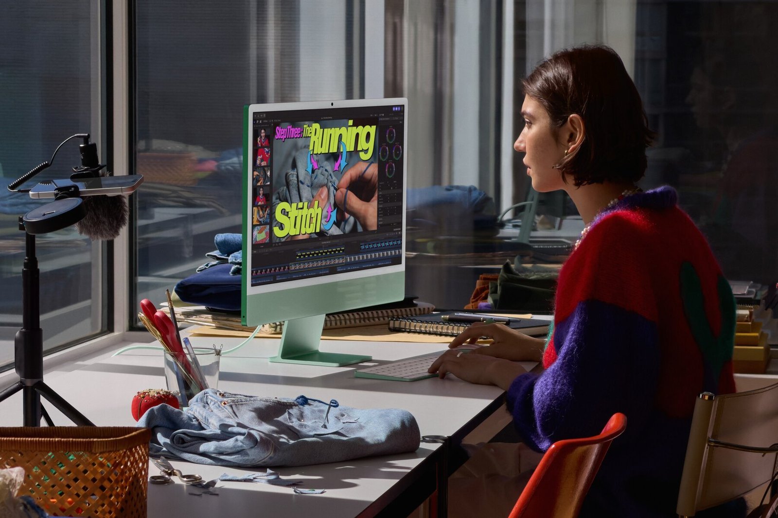A woman in a striped sweater edits video on a green iMac with M6 in a sunlit studio with a boom microphone and fabric on the desk.