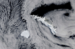 A large, blocky iceberg sits in dark ocean water just southwest of a long, narrow, snow-capped island. Thin clouds partly veil the scene.
