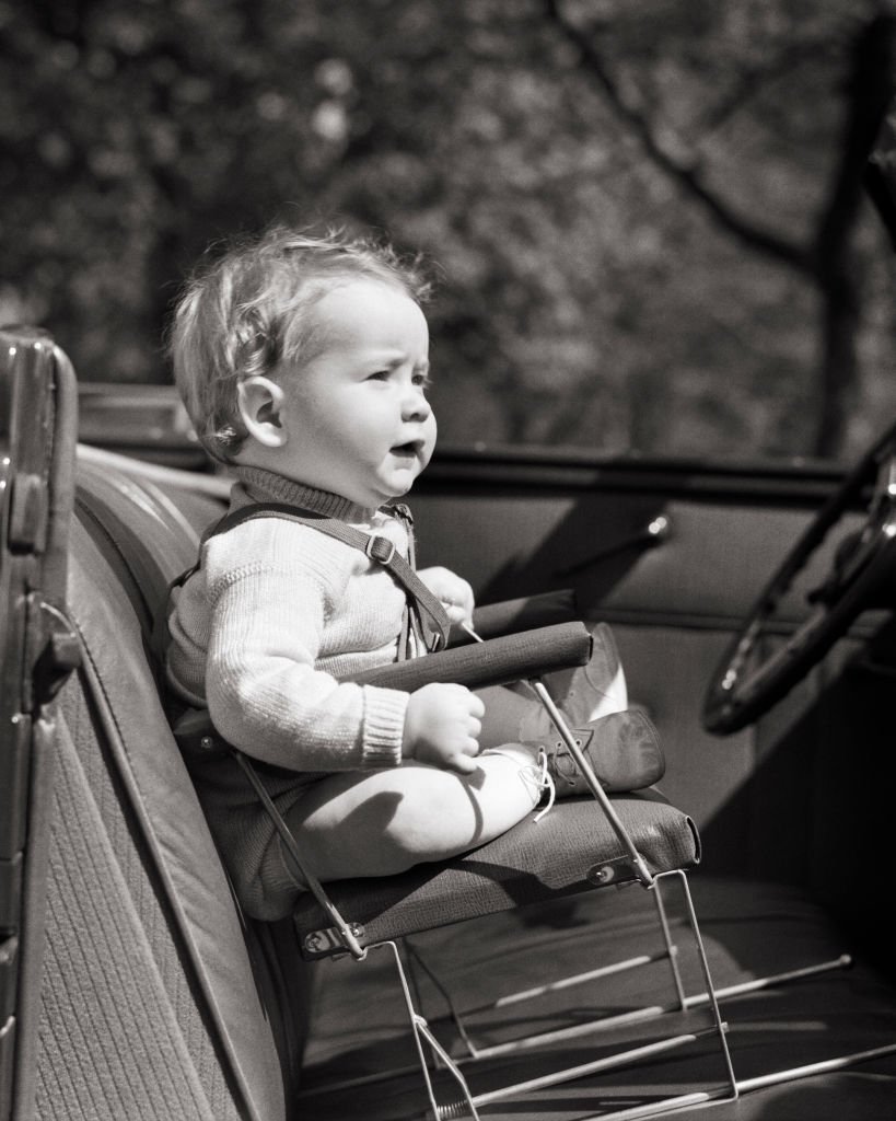 1930s 1940s baby sitting on primitive dangerous design wire child car chair no safety belt in front seat of convertible auto.