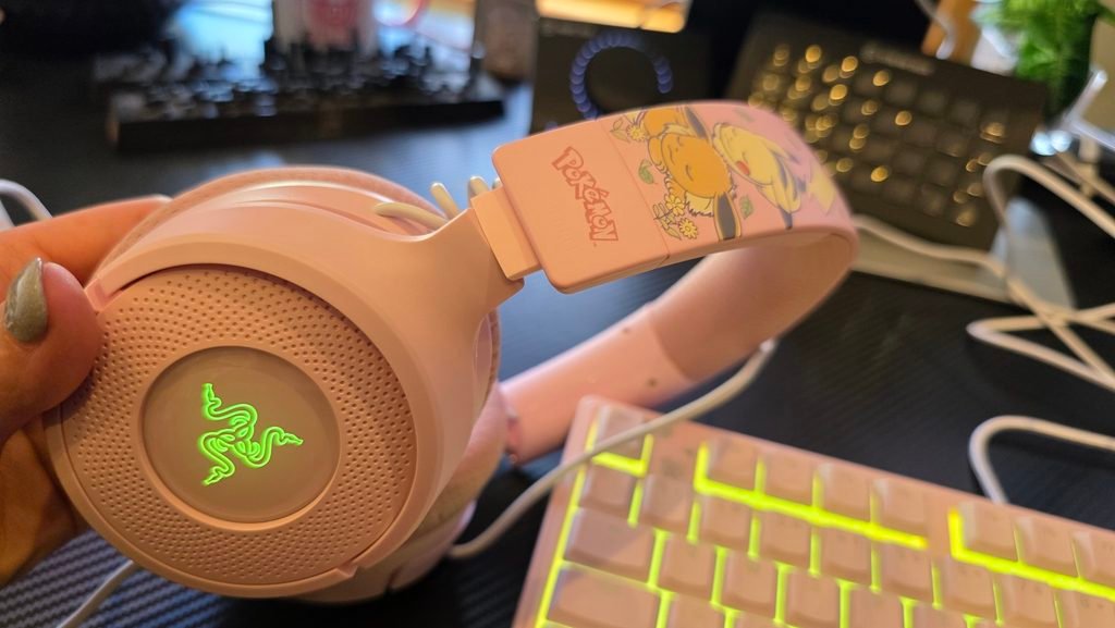 Side profile of the headset showing the Razer logo lit up, and the Pokémon Pikachu and Eevee graphics on the headband. The Pokémon Pikachu and Eevee keyboard is in the background.
