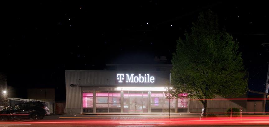 Exterior of a T-Mobile store at night under stars. The long-exposure lights of a passing car make red trails across the bottom of the photo.