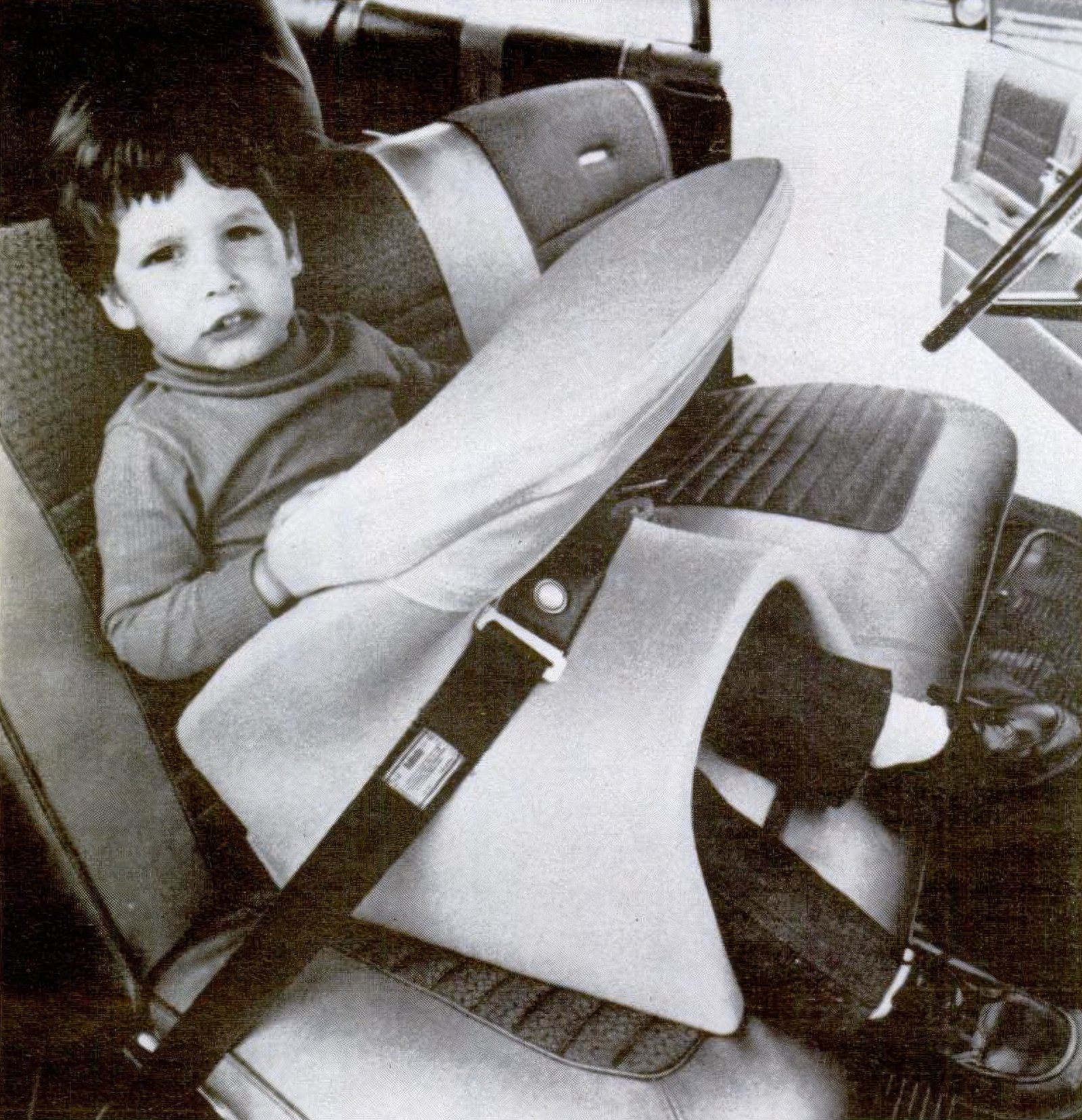 Young boy sits in Ford Tot Guard car seat in black and white photograph. The Tot Guard is white and goes in front of child and has a head impact cushion.