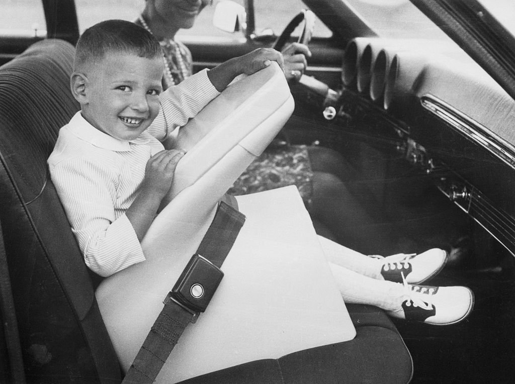 Black and white photograph of smiling young boy in vintage car seat next to smiling woman whose face is cut off.